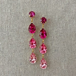 May include: A pair of dangling earrings with a gold-tone metal setting. Each earring features a round pink gemstone at the top, followed by three teardrop-shaped pink gemstones that graduate in color from dark to light. The earrings are displayed on a neutral linen background.