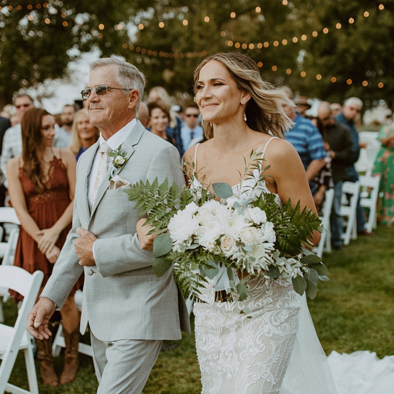 May include: A bride in a white lace wedding dress walks down the aisle with her father. She carries a bouquet of white flowers and greenery. The father wears a light gray suit and sunglasses. Guests are seated on either side.