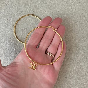 May include: Gold-colored hoop earrings. One earring features a small bird charm. The earrings are held in a hand, with rings on the fingers. The hoops are large and have a simple, elegant design.