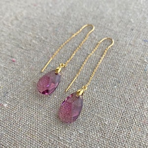 May include: A pair of gold-colored threader earrings with teardrop-shaped, faceted, purple gemstones. The earrings hang from delicate chains, and the gemstones have a gradient of color. The earrings are displayed on a textured, neutral-colored surface.