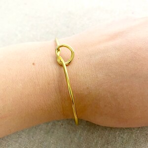 May include: A gold-coloured bracelet with a knot design. The bracelet is thin and delicate, with a simple, elegant style. The knot is the focal point, adding a unique touch to the jewellery. The bracelet is shown on a wrist.