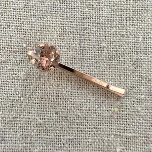 May include: A rose gold hair pin with a round, faceted, light pink gemstone at the top. The pin is lying on a textured, light gray surface. The gemstone is surrounded by small prongs.