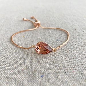 K&ouml;nnte beinhalten: Ein verstellbares Armband aus Ros&eacute;gold mit einem tropfenf&ouml;rmigen, pfirsichfarbenen Kristall. Der Kristall ist in einem filigranen Rahmen gefasst, und das Armband hat eine feine Kette mit einem Schiebeverschluss. Das Armband wird auf einer neutralen Leinenoberfl&auml;che pr&auml;sentiert.