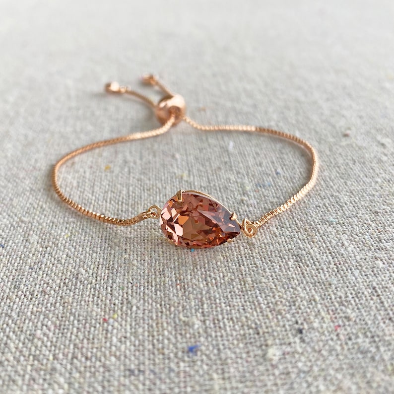 K&ouml;nnte beinhalten: Ein verstellbares Armband aus Ros&eacute;gold mit einem tropfenf&ouml;rmigen, pfirsichfarbenen Kristall. Der Kristall ist in einem filigranen Rahmen gefasst, und das Armband hat eine feine Kette mit einem Schiebeverschluss. Das Armband wird auf einer neutralen Leinenoberfl&auml;che pr&auml;sentiert.
