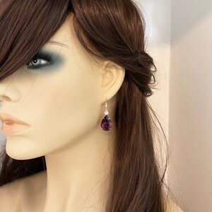 May include: A single gold-toned earring with a teardrop-shaped, deep purple gemstone. Above the purple stone is a smaller, round, clear stone. The earring is shown on a mannequin with brown hair.