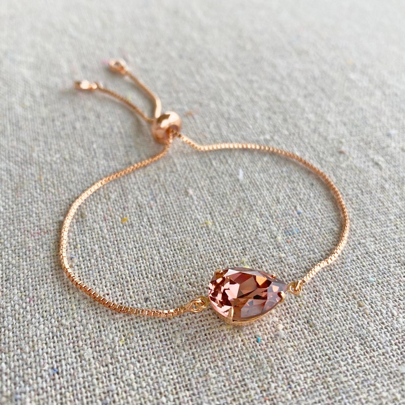 K&ouml;nnte beinhalten: Verstellbares Armband in Ros&eacute;gold mit einem tropfenf&ouml;rmigen, hellbraunen Kristall. Das Armband hat eine feine Kette und einen Schiebeverschluss f&uuml;r eine individuelle Passform. Der Kristall ist das Hauptmerkmal des Armbands.