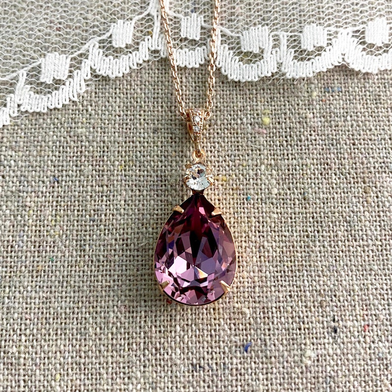 Puede incluir: Un collar de oro rosa con un colgante grande en forma de l&aacute;grima de color rosa oscuro. El colgante est&aacute; facetado y tiene un cristal claro redondo m&aacute;s peque&ntilde;o encima. La cadena es delicada, sobre un fondo de tela neutra texturizada.