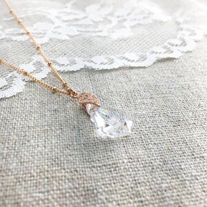 May include: A rose gold necklace featuring a clear, teardrop-shaped pendant. The pendant is faceted, reflecting light. The delicate chain has small beaded details. The necklace is displayed on a textured, neutral surface, with a white lace trim.