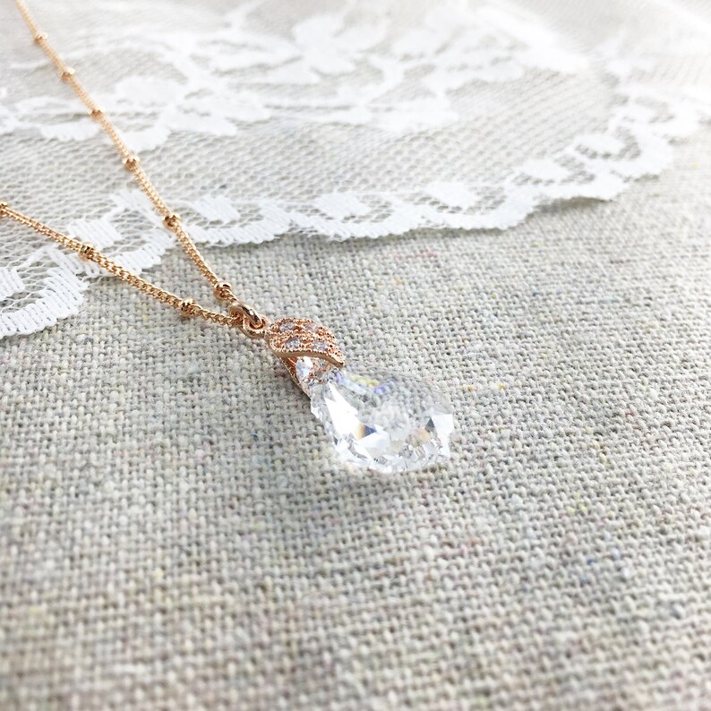 May include: A rose gold necklace featuring a clear, teardrop-shaped pendant. The pendant is faceted, reflecting light. The delicate chain has small beaded details. The necklace is displayed on a textured, neutral surface, with a white lace trim.