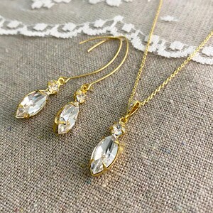 May include: A gold-toned jewelry set featuring a necklace and a pair of earrings. The set includes a pendant necklace and drop earrings, each with a large, clear marquise-cut crystal and smaller accent stones. The jewelry is displayed on a neutral-toned surface.