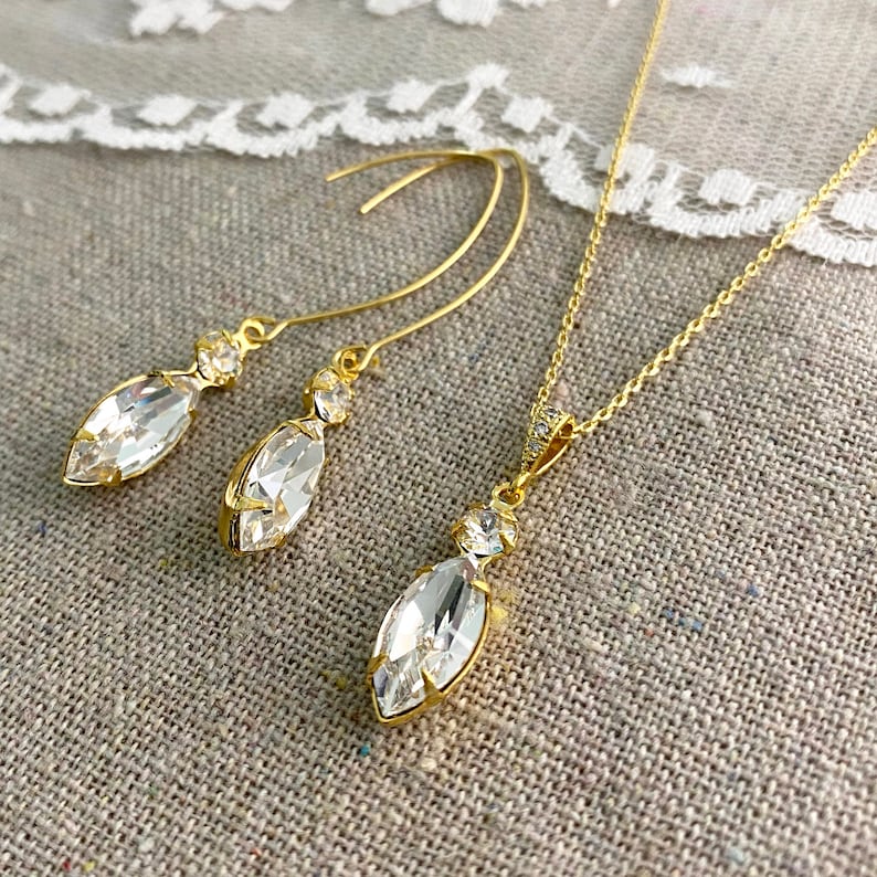 May include: A gold-toned jewelry set featuring a necklace and a pair of earrings. The set includes a pendant necklace and drop earrings, each with a large, clear marquise-cut crystal and smaller accent stones. The jewelry is displayed on a neutral-toned surface.