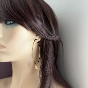 May include: Gold-colored hoop earrings with a small bird charm. The earrings are large and circular, with a simple, elegant design. The bird charm is a delicate, detailed silhouette.