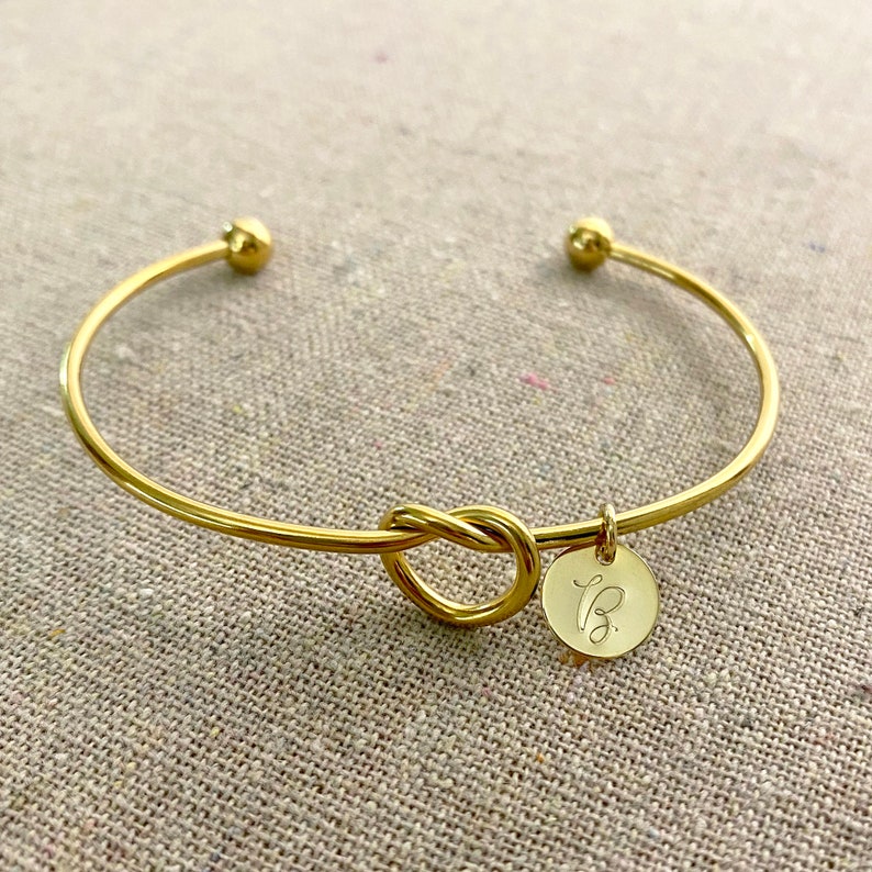 May include: A gold-coloured bangle bracelet featuring a knot design at the centre. It has a small, circular charm engraved with the letter "B". The bracelet is made of a shiny, metallic material.
