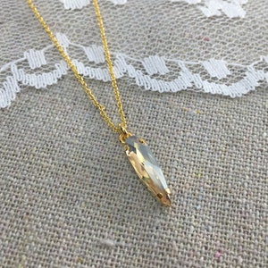 Puede incluir: Collar dorado con un colgante de cristal alargado de color claro. El cristal tiene forma puntiaguda y está engastado en metal dorado. La cadena también es dorada y delicada. El cristal mide unos 3 cm de largo.