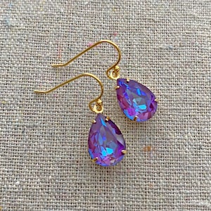 May include: A pair of gold-colored drop earrings with teardrop-shaped, iridescent purple and blue crystals. The earrings are displayed on a textured, neutral-colored surface.