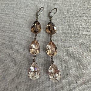May include: A pair of dangle earrings with three teardrop-shaped crystals in shades of brown and champagne. The crystals are connected by small metal links and have silver-colored ear wires.
