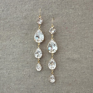 May include: Elegant gold-toned drop earrings featuring clear, sparkling crystals. Each earring has a top round crystal, followed by three descending pear-shaped crystals. The earrings are suspended from a simple hook.
