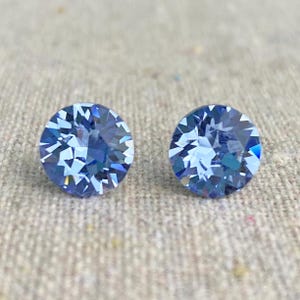May include: Two round, faceted, light blue gemstones. The gems have a brilliant cut, reflecting light with multiple facets. The earrings are set against a neutral, textured background, highlighting their color and sparkle. These are likely stud earrings.