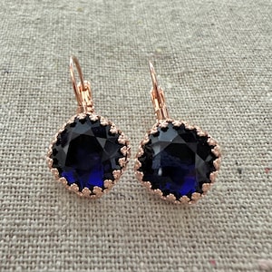 May include: A pair of rose gold leverback earrings with large, square, faceted, dark blue gemstones. The gemstones are surrounded by a decorative, scalloped edge. The earrings are displayed on a neutral, textured surface.