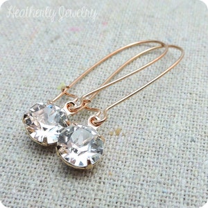 May include: Rose gold-colored earrings with clear, round crystal pendants. The earrings have a simple, elegant design with a long, curved wire that goes through the ear. The brand name "Heatherly Jewelry" is visible in the top left corner.