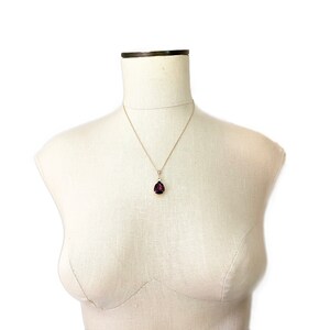 May include: A gold-toned necklace with a pear-shaped, deep purple gemstone pendant. The pendant is suspended from a delicate chain and features a small, clear accent stone above the main gem. The necklace is displayed on a mannequin.