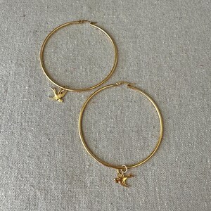 May include: A pair of gold-colored hoop earrings. Each earring has a small, gold-colored bird charm dangling from the bottom. The earrings are displayed on a neutral-colored, textured surface.