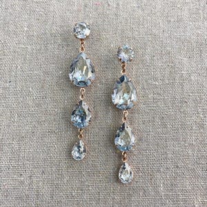May include: A pair of elegant drop earrings featuring light blue teardrop-shaped crystals and smaller round crystals. The earrings are connected by delicate gold-colored chains, creating a layered effect. The earrings are displayed on a neutral-colored fabric.
