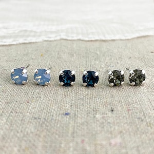 May include: Six pairs of stud earrings with round, faceted gemstones in shades of blue and gray. The earrings are set in silver-colored metal. The earrings are displayed on a neutral linen surface.