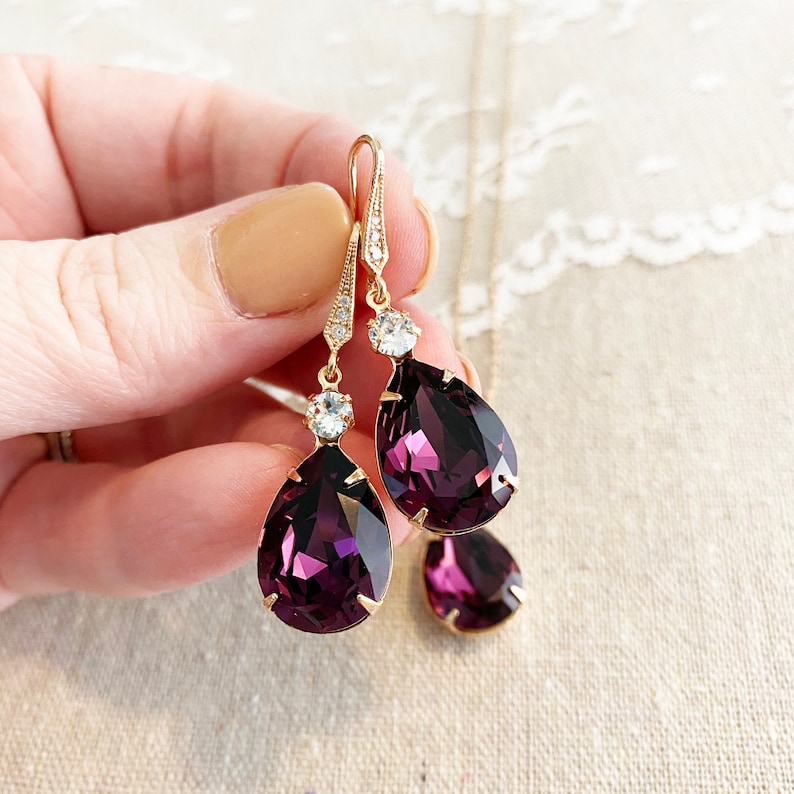May include: Gold-toned drop earrings with large, deep purple teardrop-shaped crystals. The earrings feature smaller, clear crystals and a delicate design. A matching pendant with a single teardrop crystal is also visible.