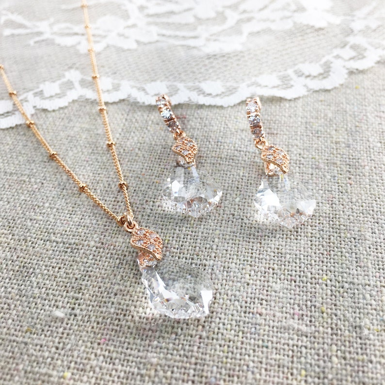 May include: A rose gold necklace and earring set. The necklace has a fine chain with a clear, faceted crystal pendant. The earrings mirror the design, with clear crystals and rose gold detailing. The jewellery set is presented on a neutral fabric background.