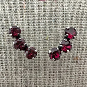 May include: A pair of silver-toned stud earrings, each featuring three round, deep red gemstones. The gemstones are set in a curved arrangement, creating a delicate and elegant design. The earrings are displayed on a textured, neutral-toned surface.