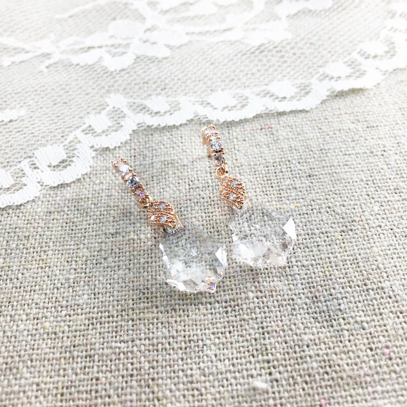 May include: Rose gold drop earrings with clear, faceted crystal pendants. The earrings have a delicate design with small, sparkling crystals along the top. The crystals are set in a geometric pattern, adding elegance to the jewellery.
