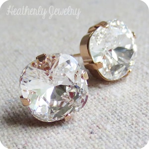May include: A pair of clear, faceted crystal stud earrings with rose gold settings. The earrings are round and sparkling, with the Heatherly Jewelry logo in the top left corner.