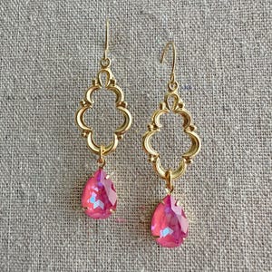 May include: A pair of gold-colored drop earrings. Each earring features an ornate, openwork design with a teardrop-shaped, pink crystal pendant. The earrings are displayed against a neutral, textured background.