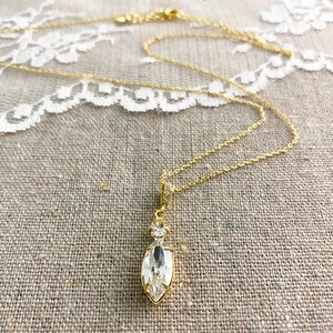 May include: A gold-colored necklace with a pendant featuring a large, clear, marquise-cut crystal. The pendant is accented with smaller, round crystals. The necklace is displayed on a textured, neutral-colored surface.