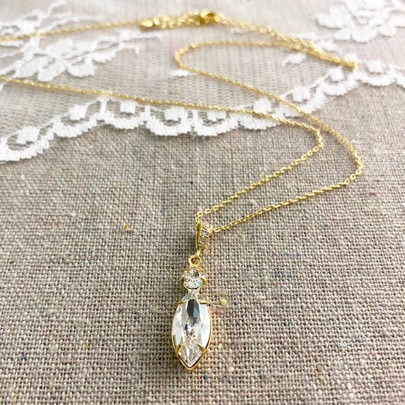 May include: A gold-colored necklace with a pendant featuring a large, clear, marquise-cut crystal. The pendant is accented with smaller, round crystals. The necklace is displayed on a textured, neutral-colored surface.