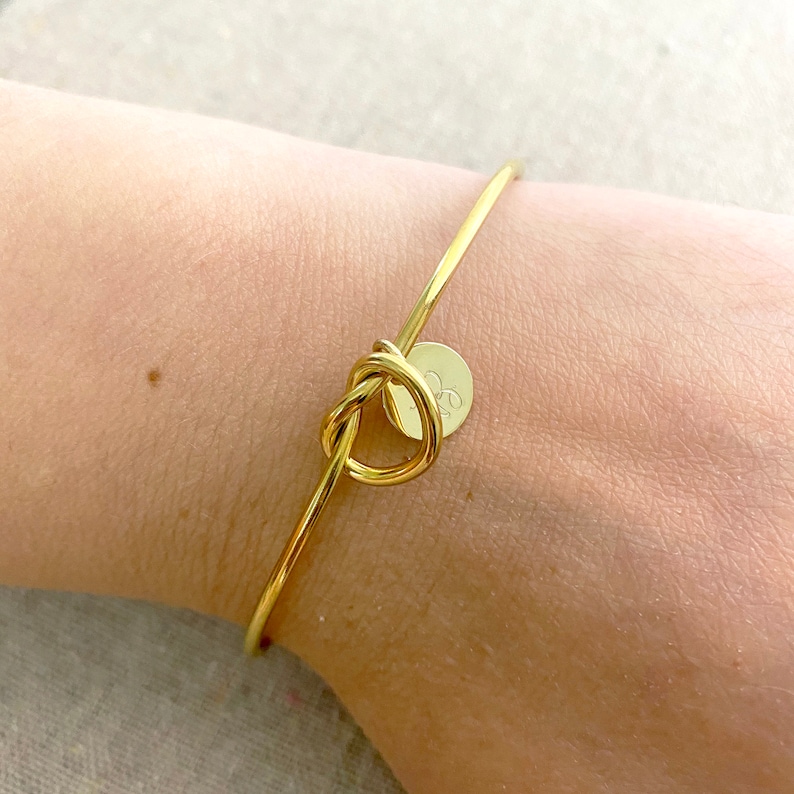 May include: A gold bangle bracelet with a knot design at the centre. A small, round charm with an initial is attached. The bracelet is worn on a wrist.