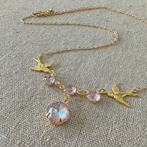 May include: A gold-toned necklace featuring a delicate chain and a series of iridescent pink crystals. The necklace is adorned with two gold bird charms, adding a touch of nature-inspired elegance to the jewelry.