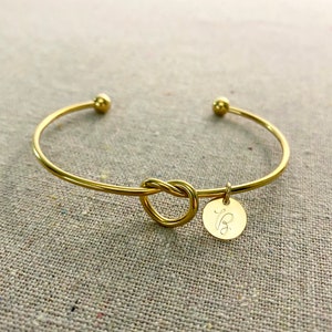 May include: Gold-coloured bangle bracelet featuring a knot design and a small, round charm. The charm has an engraved letter. The bracelet has small ball accents on each end.