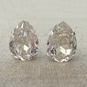 May include: Two clear, teardrop-shaped earrings with intricate facets, reflecting light. The earrings are set against a neutral, textured background, highlighting their sparkling appearance. These earrings are a classic jewelry item.