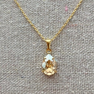 May include: A gold-toned necklace featuring a pear-shaped pendant with a light amber-colored crystal. The pendant is suspended from a delicate chain, creating an elegant and classic look. The necklace is displayed on a neutral-toned fabric.