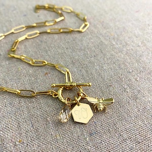 May include: Gold-colored necklace with a toggle clasp and a chain link design. The necklace features three charms: a clear crystal, a gold-colored hexagon with the letter "W", and a bee charm.