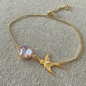 May include: Gold-toned adjustable bracelet featuring a large, iridescent, light pink crystal and a gold bird charm. The bracelet has a delicate chain and a sliding bead for adjustment. The bird charm is a stylized swallow in flight.
