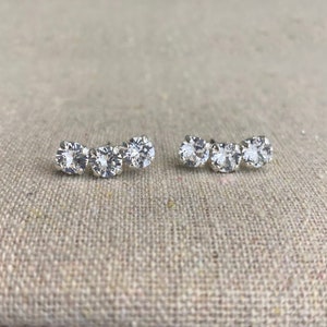 May include: A pair of silver-toned stud earrings, each featuring three clear, round-cut cubic zirconia stones in a curved row. The earrings are displayed on a neutral-toned, textured surface.