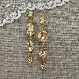 May include: A pair of elegant, gold-toned drop earrings. Each earring features a small, round crystal at the top, followed by three teardrop-shaped crystals of varying sizes. The crystals have a champagne hue and are connected by delicate gold chains.