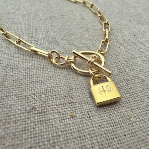 May include: Gold-colored necklace with a padlock pendant. The necklace features a toggle clasp and a rectangular padlock charm with the initials "HQ" engraved on it. The chain links are rectangular.