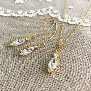 May include: A gold-toned jewelry set featuring a necklace and earrings. The necklace has a delicate chain with a marquise-shaped pendant. The earrings are long, curved, and also feature marquise-shaped crystals. The crystals are clear and faceted, reflecting light.