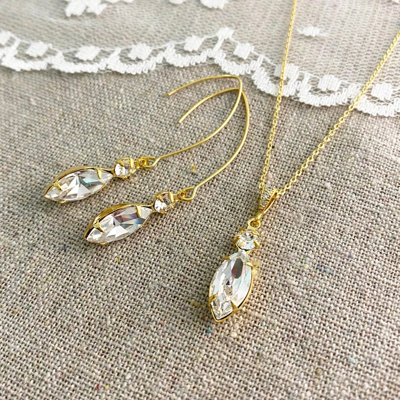 May include: A gold-toned jewelry set featuring a necklace and earrings. The necklace has a delicate chain with a marquise-shaped pendant. The earrings are long, curved, and also feature marquise-shaped crystals. The crystals are clear and faceted, reflecting light.