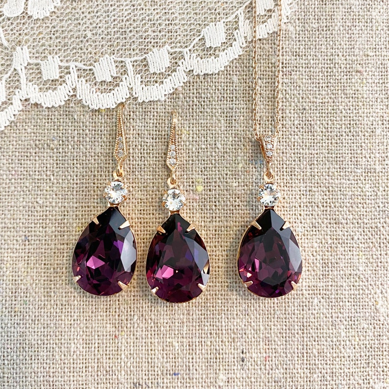 May include: A jewelry set featuring a gold-toned necklace and matching earrings. The earrings and pendant have a teardrop-shaped, deep purple gemstone, topped with a clear, round stone. The earrings are drop style.