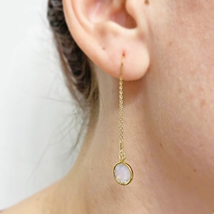 May include: A gold-colored earring with a delicate chain and a round, light-colored gemstone. The earring is hanging from the ear. The gemstone has a subtle, iridescent quality.